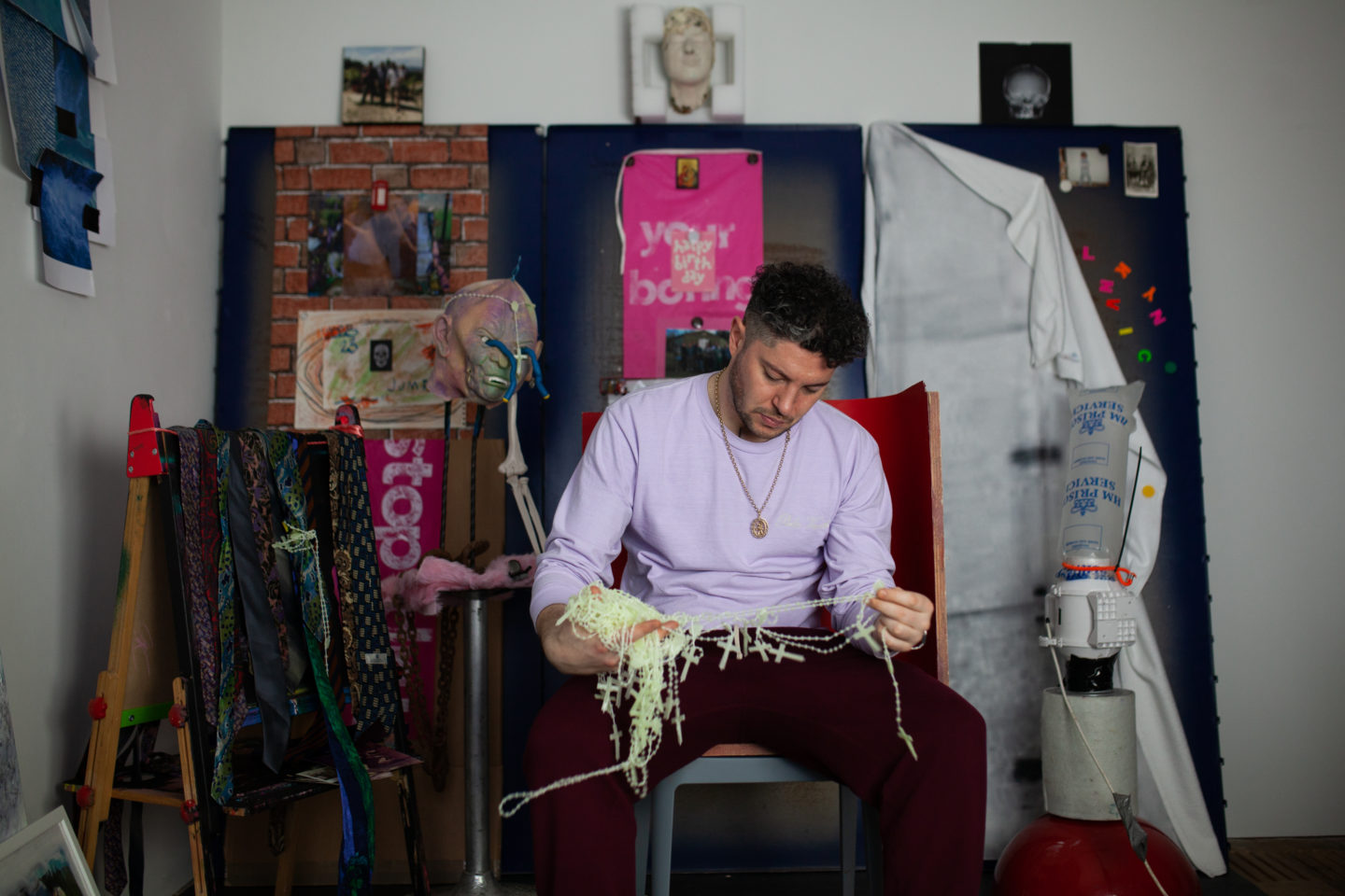 A man in a lilac jumper sits in his art studio looking at a pile of glow in the dark rosaries