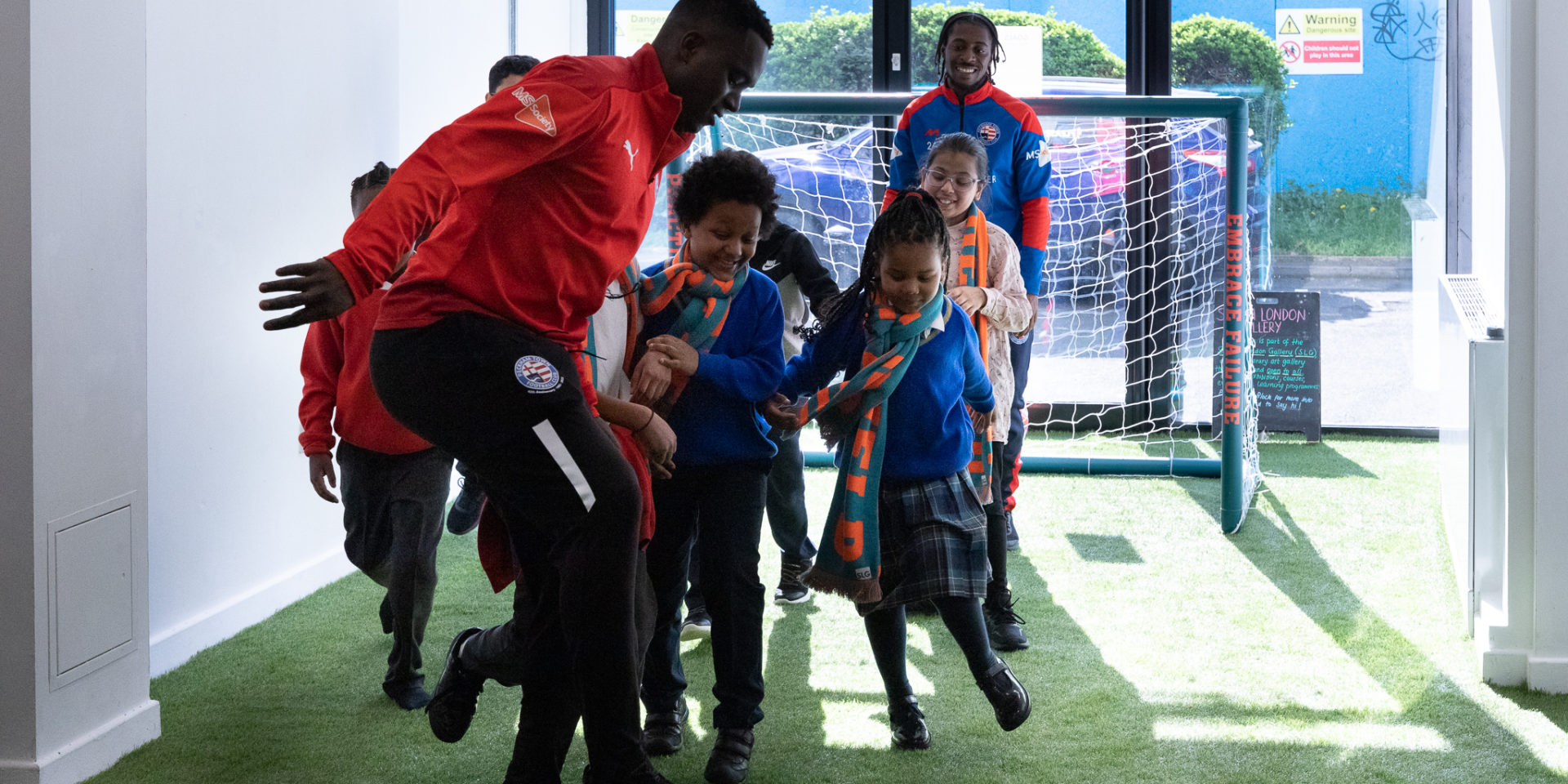 Children and adults play football in a bright room. The room is carpeted with astro turf and there is a goal post at one end.