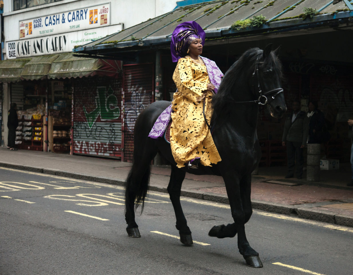 A woman wearing traditional Nigerian clothes rides a horse through the streets of Peckham, London