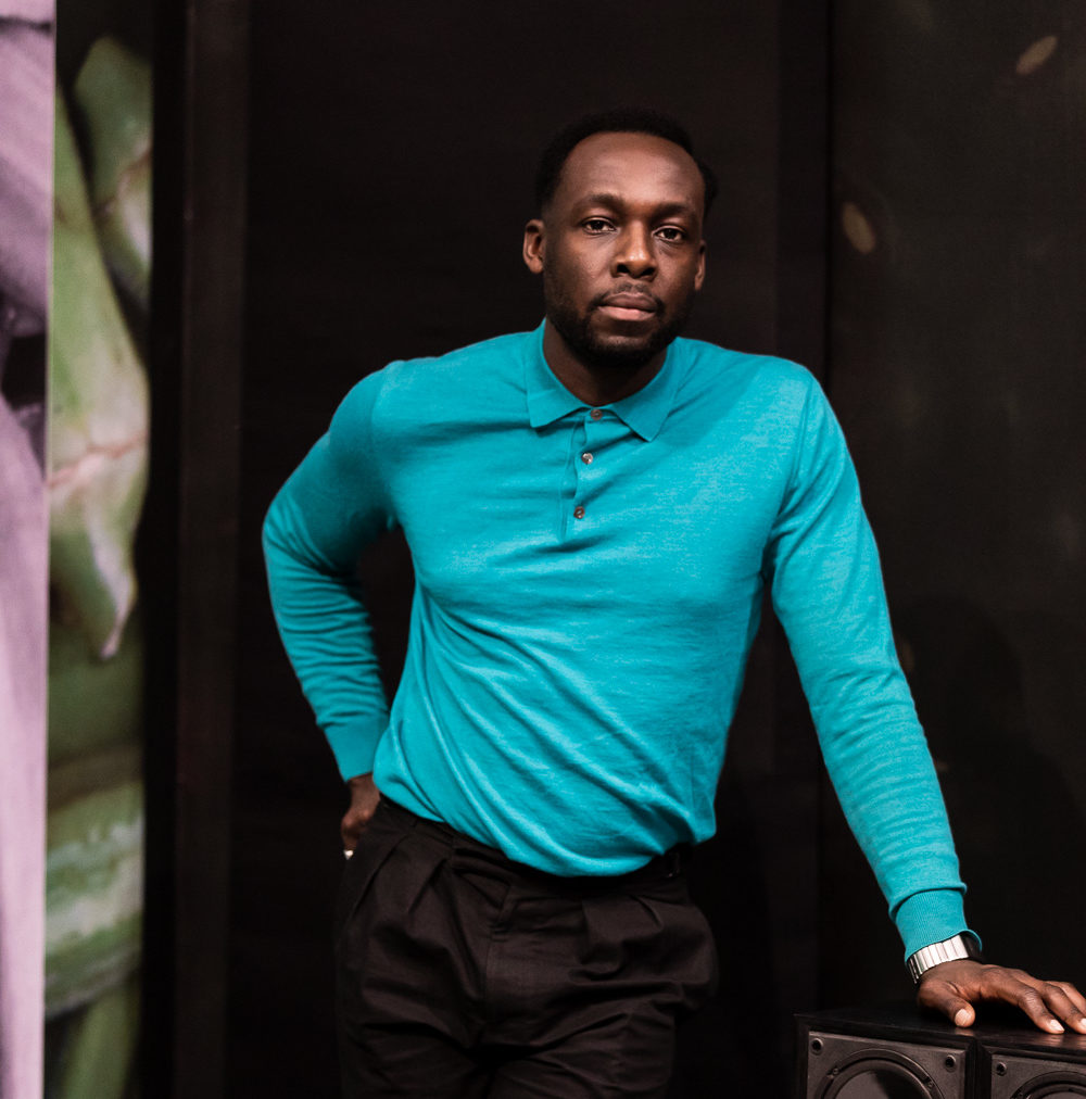 A man wearing a bright, teal coloured top stands leaning against a table.