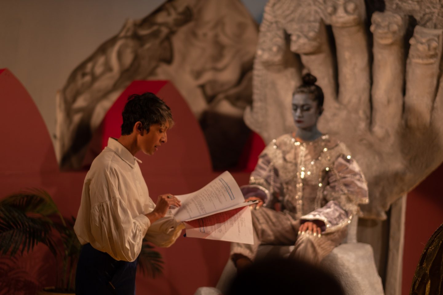 A woman wearing a white shirt and reading a script stands in the middle of a film set. Behind her, an actor painted all in blue wearing a silver costume sits on a large grey throne.