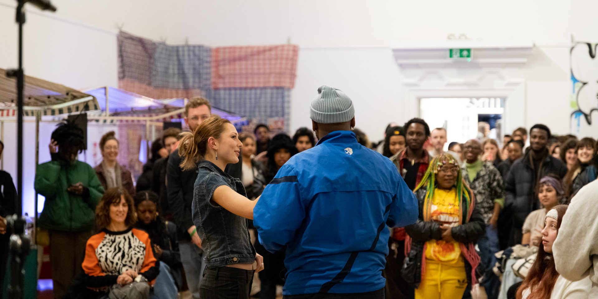 Two people stand in front of a crowd in a large, bright gallery space. The woman holds out a microphone to a man wearing a blue top, who is speaking to the crowd.