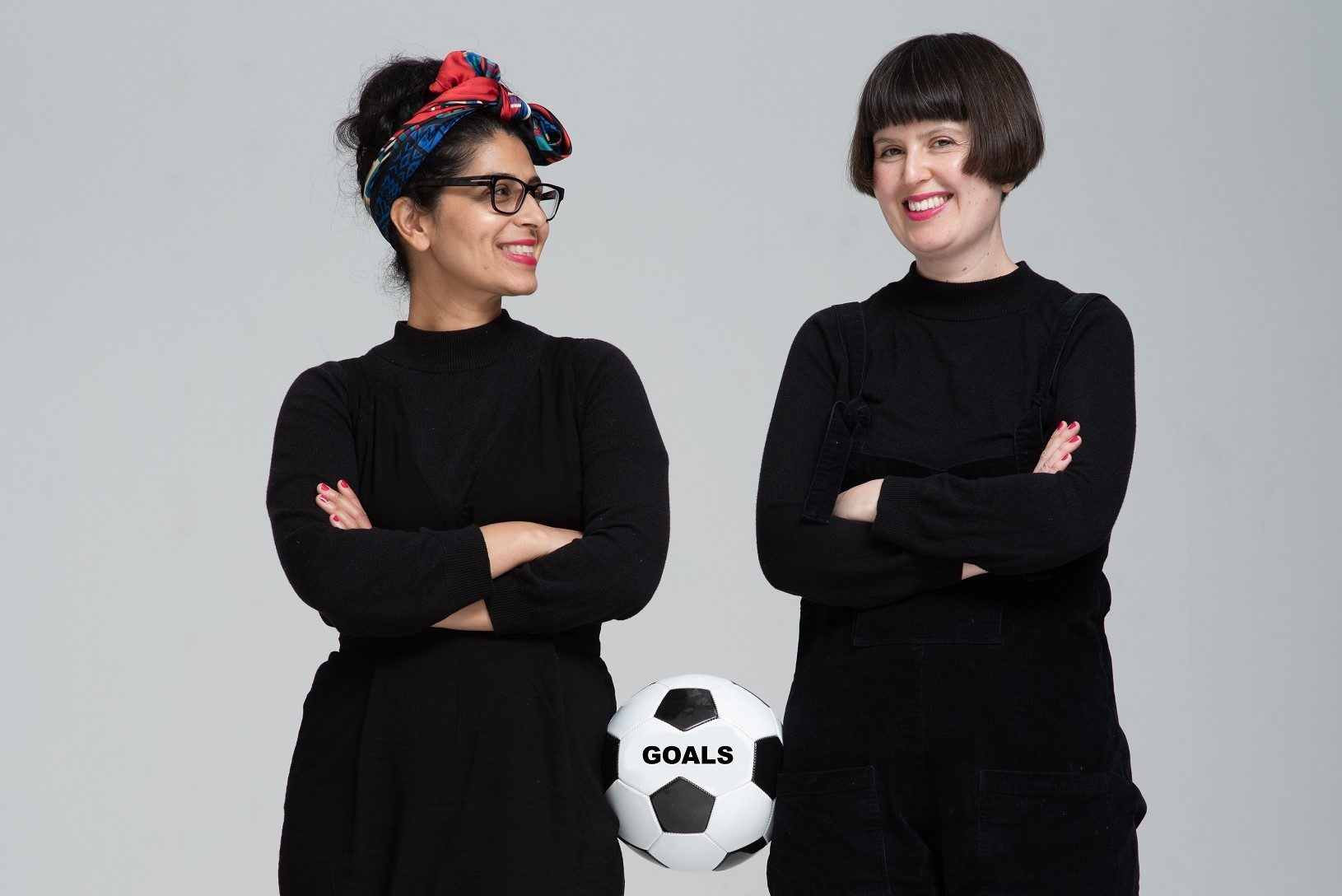 Two women wearing black stand with their arms crossed. There is a football balanced on their hips between them.