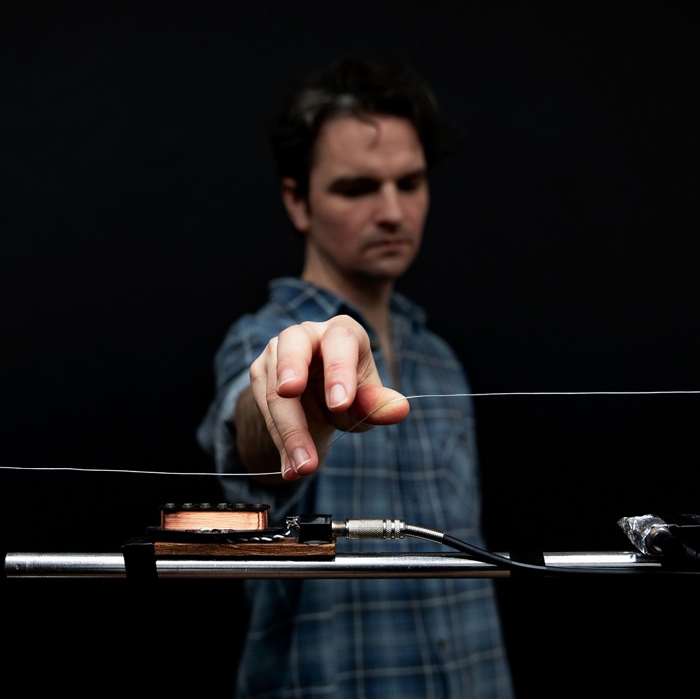 A man stands with his hand out, reaching towards the camera. His hand hovers over a handmade musical instrument.