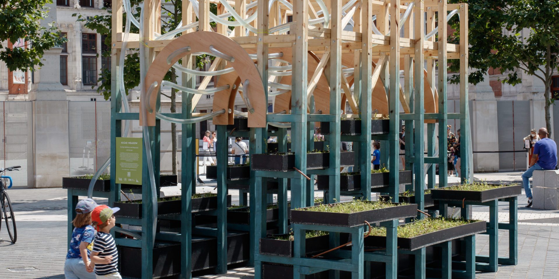A structure with children running about it. The structure is made from wood and has plastic pipes running through it. There are trays with plants around the outside.