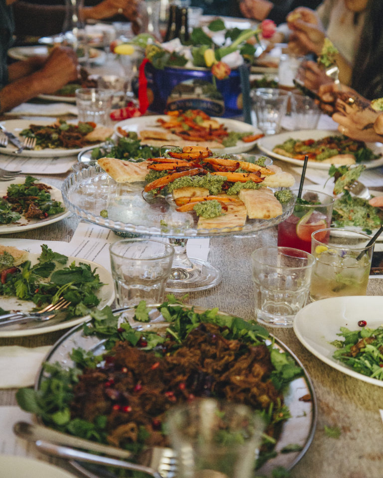 A table filled with plates of food in the background there are people sat around the table