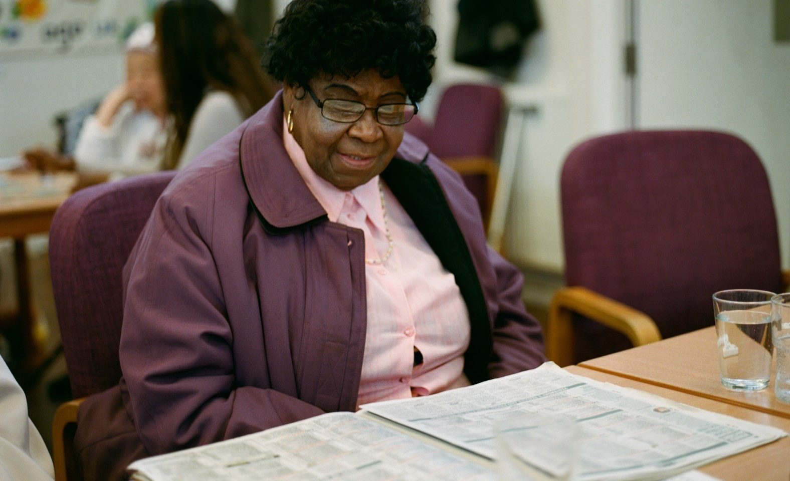 A woman sits at a wooden table. She is wearing a purple jacket and is reading a newspaper laid out on the table in front of her.