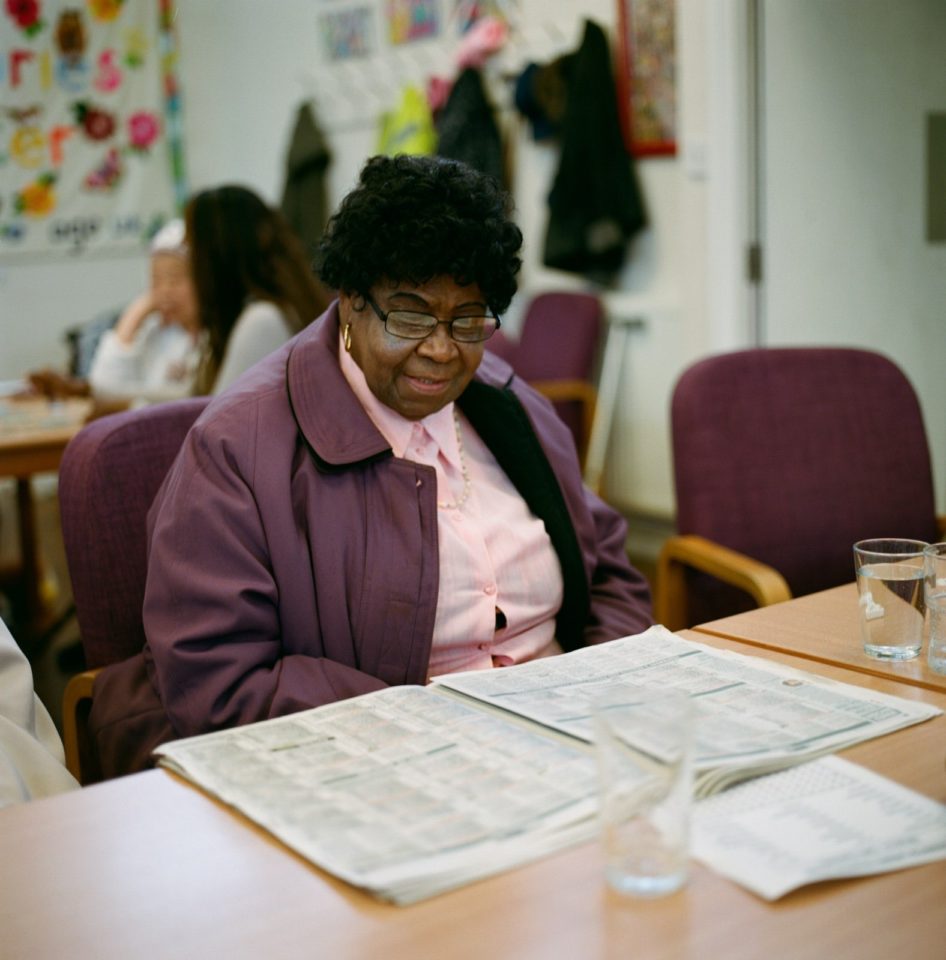 A woman sits at a wooden table. She is wearing a purple jacket and is reading a newspaper laid out on the table in front of her.