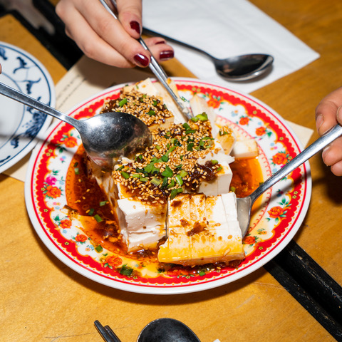 Hands holding spoons reach out to a plate on a table. The colourful plate has tofu on it.