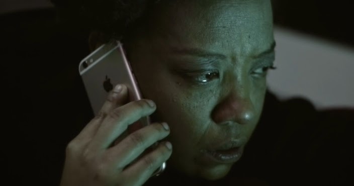 A woman sits in a dark room with her phone held to her face. Her eyes are welling up with tears.