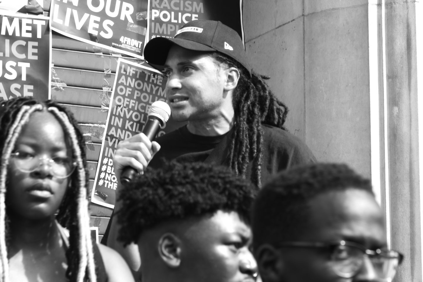 A man stands in a crowd and holds a microphone. He is wearing a black cap.