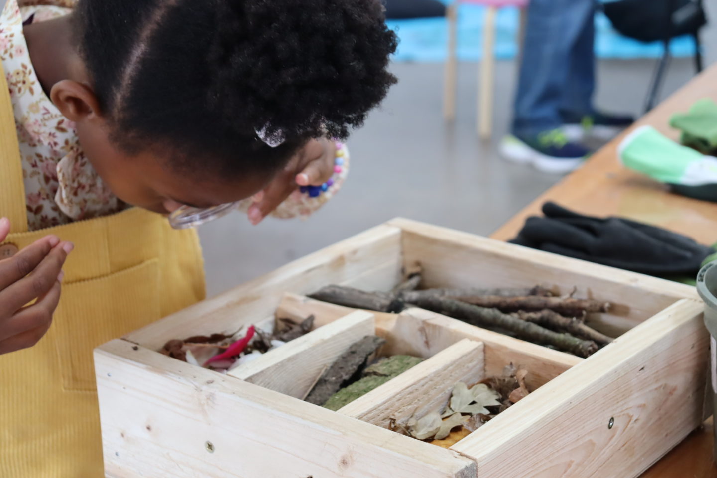 A small child wearing yellow holds a magnifying glass to their face. They are looking at a wooden structure with sticks and leaves in it.