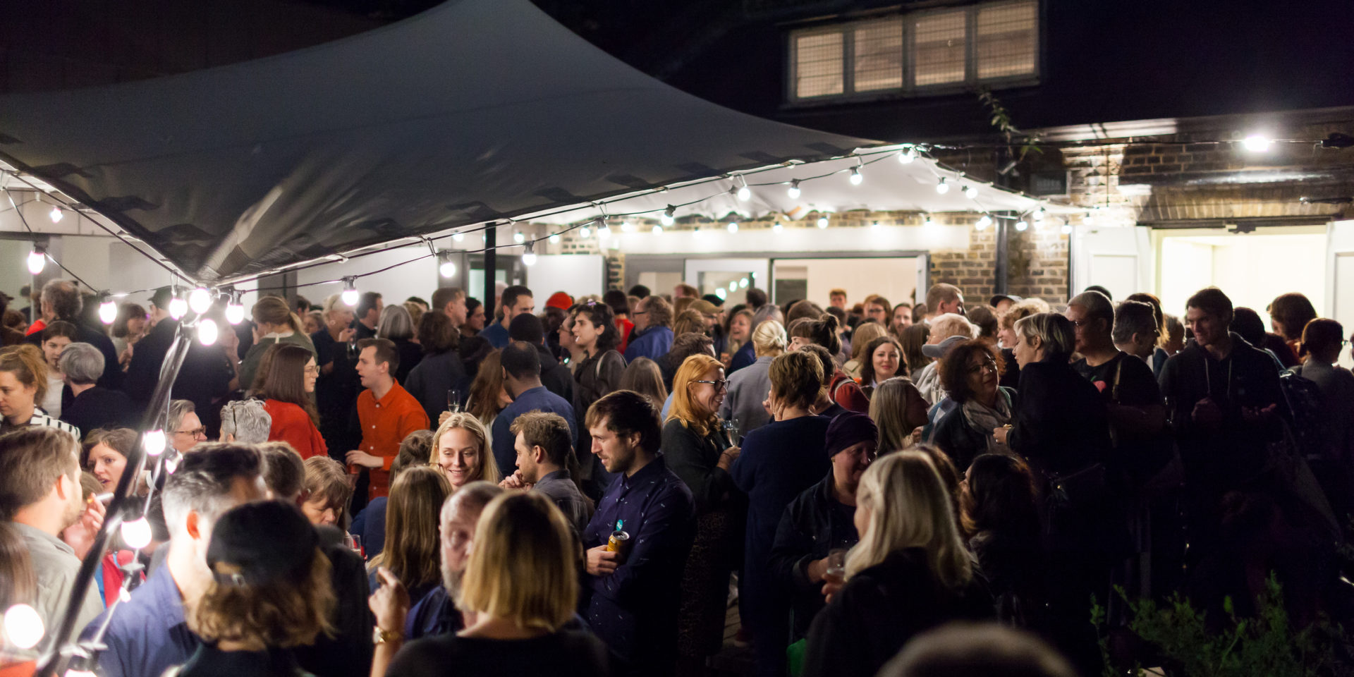 A crowd of people stand outside at nighttime. They are drinking and chatting. There is a stretch tent above them and some lights.