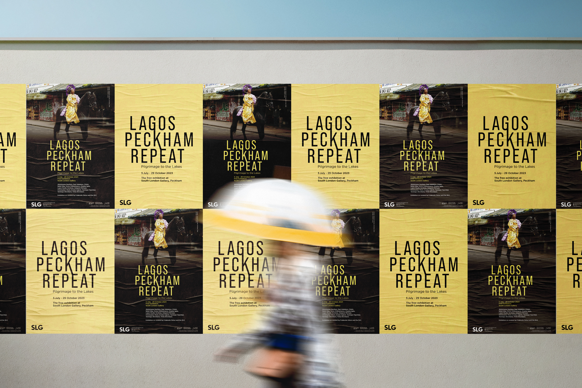 A person holding an umbrella walks past a wall of posters. The posters advertise the exhibition: Lagos, Peckham, Repeat at the South London Gallery.