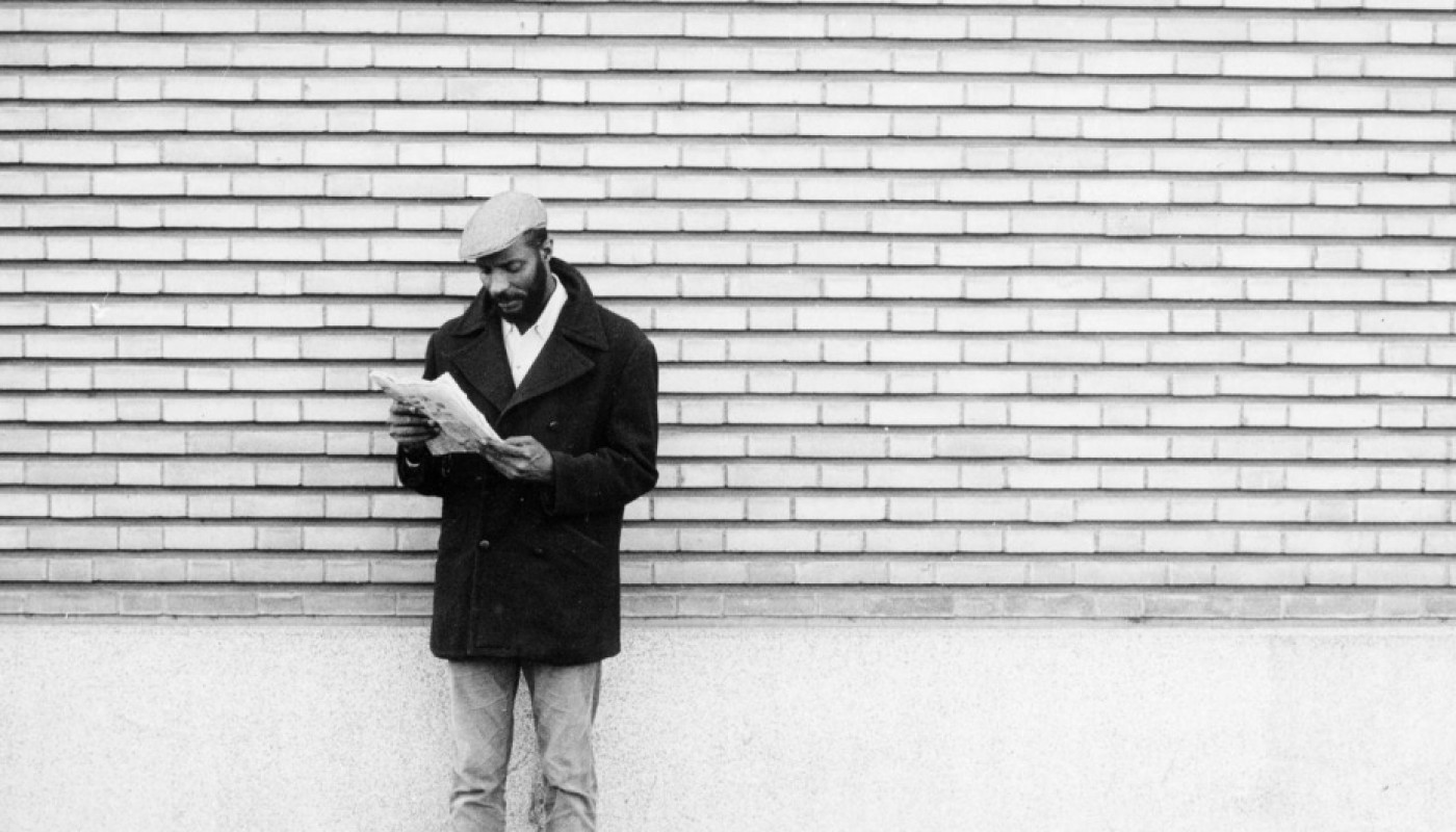 A man wearing a hat and reading a newspaper stands against a white wall.