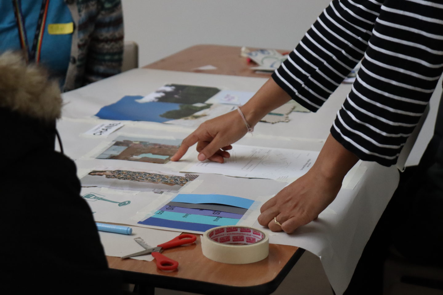 A photo of a table with lots of pages and images laid out on it. A person wearing a striped top leans on the table and points at one of the pages.