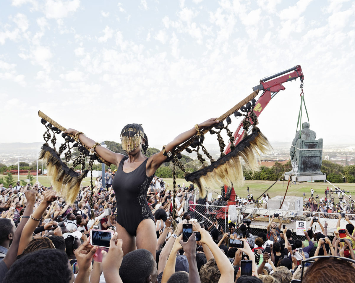 A woman with her arms aloft standing in a crowd with her back to a crane removing a statue of Cecil Rhodes