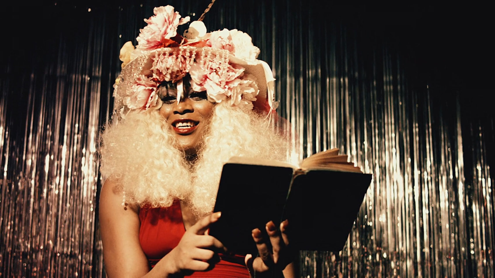 A still from a film with transgender rights activist Martha P Johnson wearing a red dress and holding a book on a stage with a backdrop of silver sparkly curtains