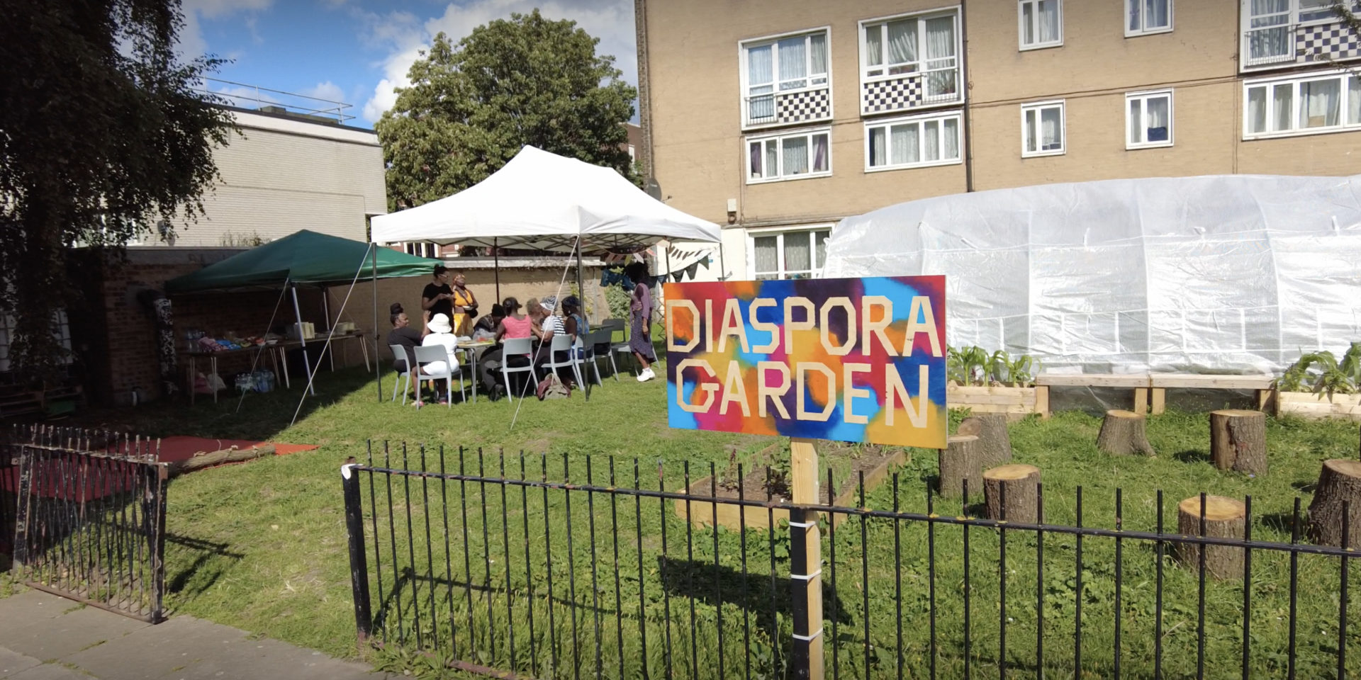 A green park space outside of a block of flats. People sit under a gazebo to shelter from the sun. A colourful sign reads: Diaspora Garden.