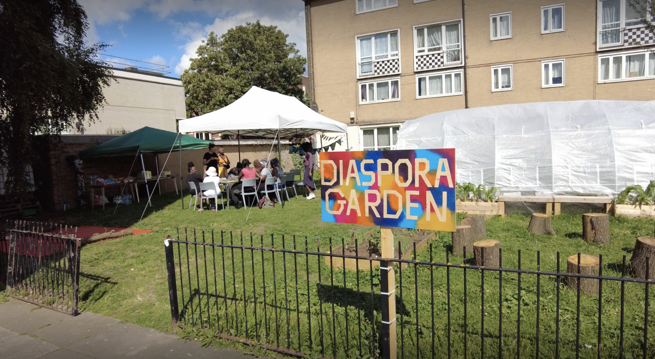 A green park space outside of a block of flats. People sit under a gazebo to shelter from the sun. A colourful sign reads: Diaspora Garden.