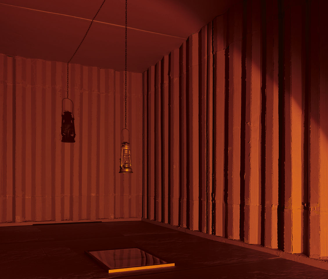 A dark container with orange light. The walls are made of cardboard and a lantern hangs in the middle above a glass frame.