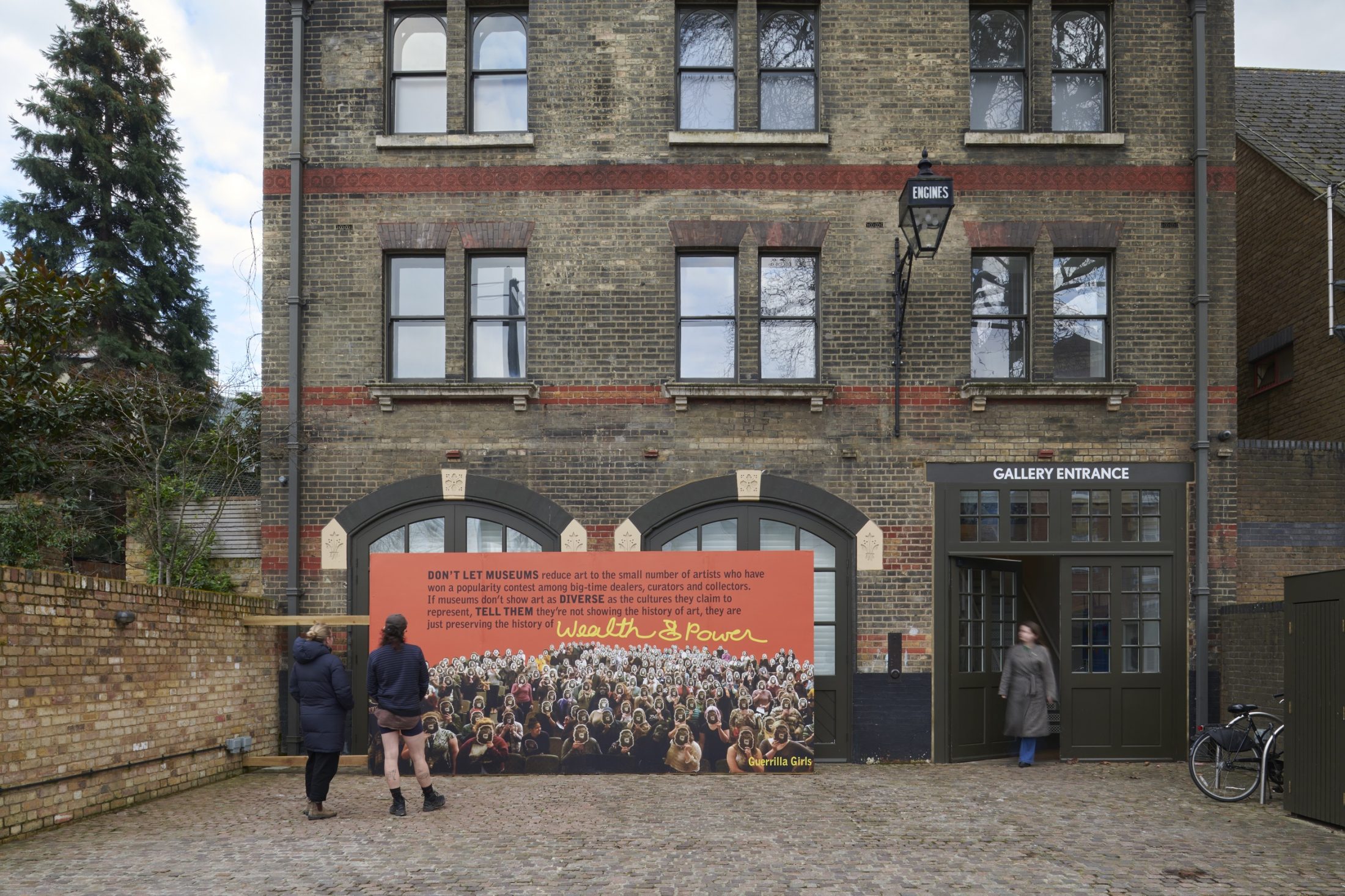 A billboard out of an old Fire Station building in Peckham. People stand on the street and look at it. The billboard displays an artwork by artist collective Guerilla Girls.