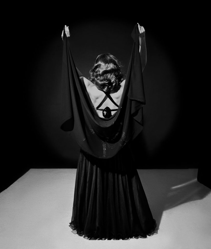 A black and white photo of a person wearing a dramatic long black dress and lifting their hands above their heads.