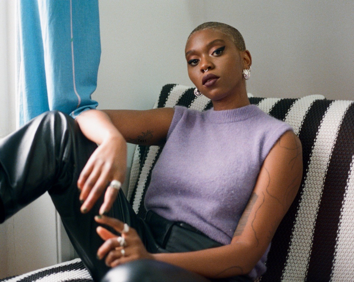 A person sits on a stripy black and white chair. They are wearing a purple top and black leather trousers.