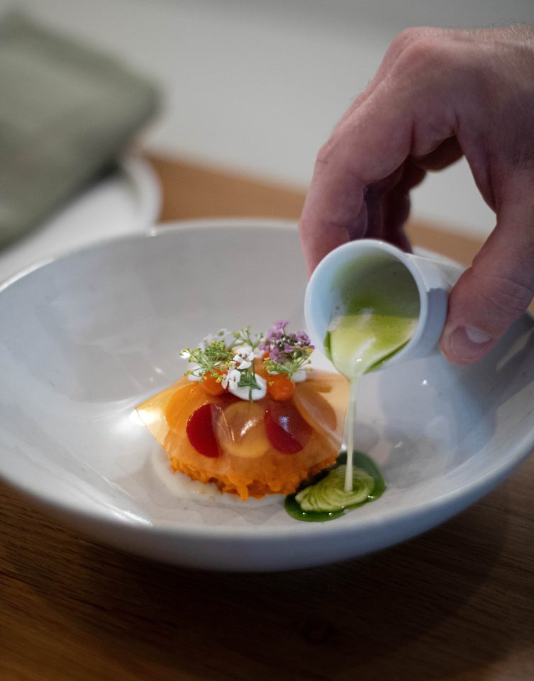 A person pours sauce over a small plate dish. The meal is part of a fine dining supper club menu.