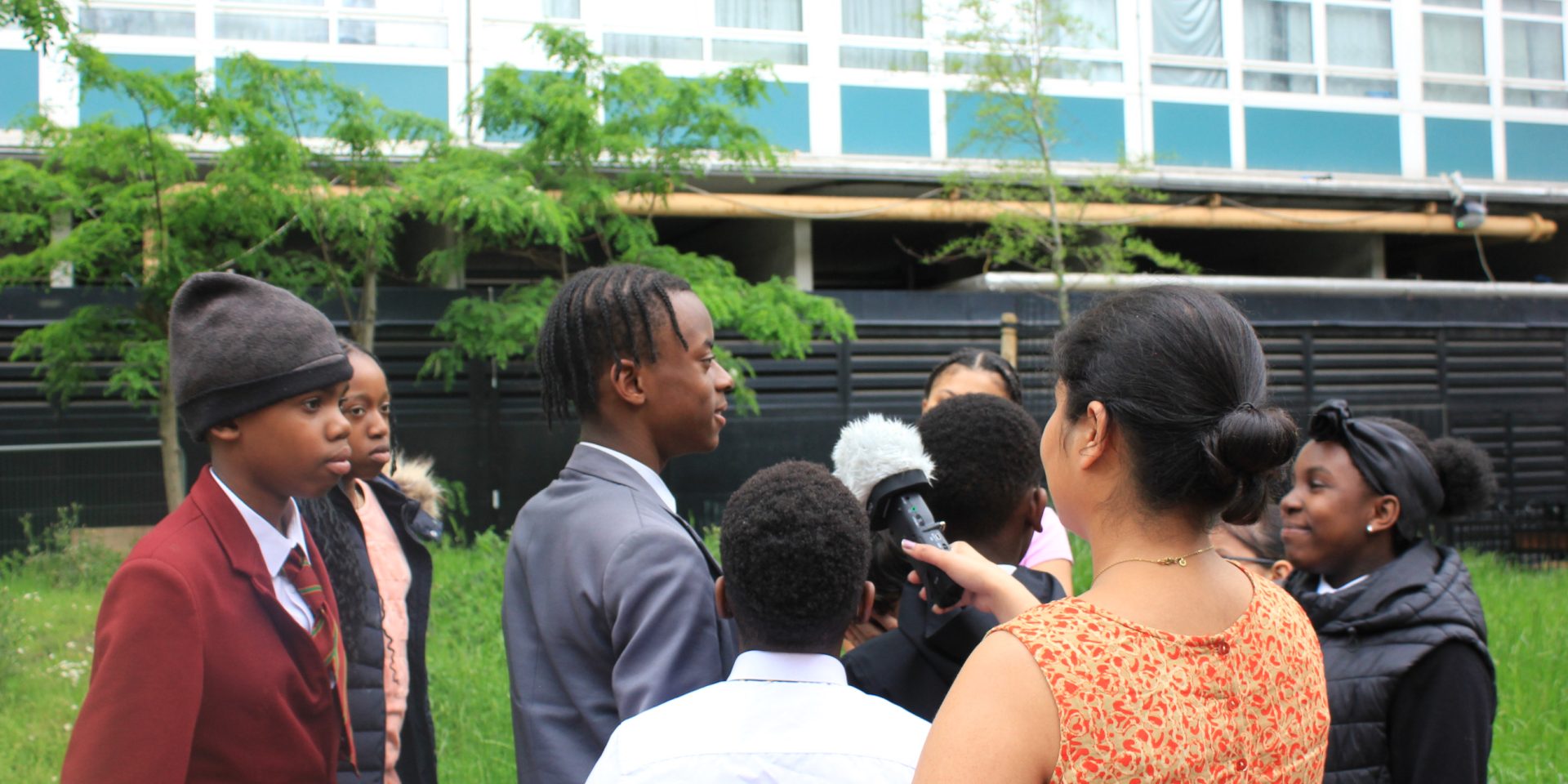 A group of young people stand around outside a building and speak to a microphone. The mic is being held by one half of artist duo, Kneed.
