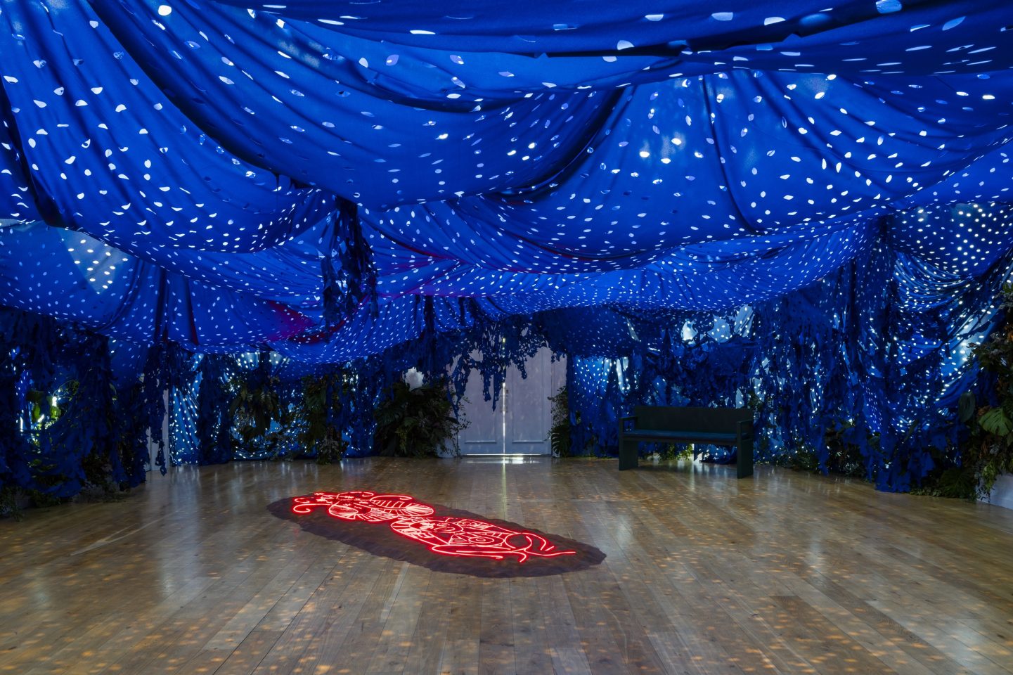 A large art gallery space with an immersive installation. The art work has blue tarp hanging from the ceiling and a red neon work on the ground.