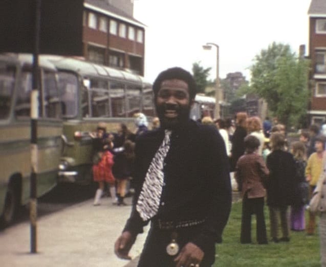 A man stands laughing and looking to camera. He is wearing all black and a tie. Behind him is a large group of children.