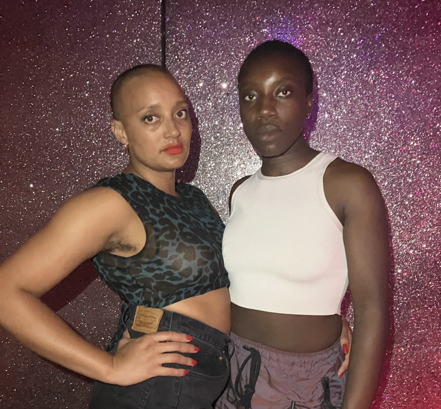 Two people stand in front of a glittery pink wall and look to camera.