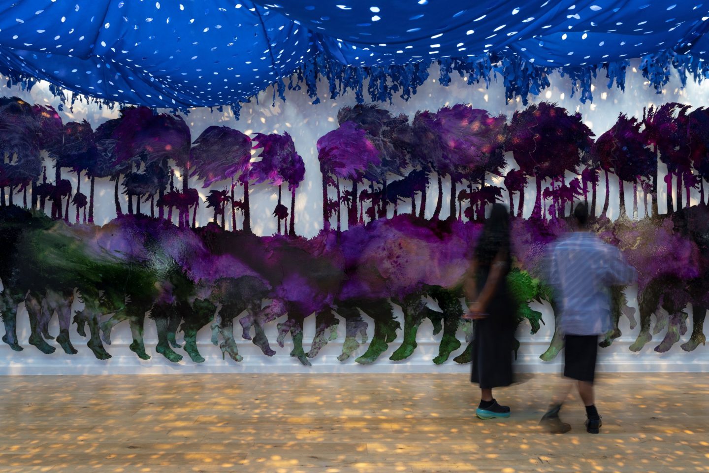An immersive art installation of an aluminum painted mythical creature. Blue tarp hangs from the ceiling. Two people are looking at the creature.