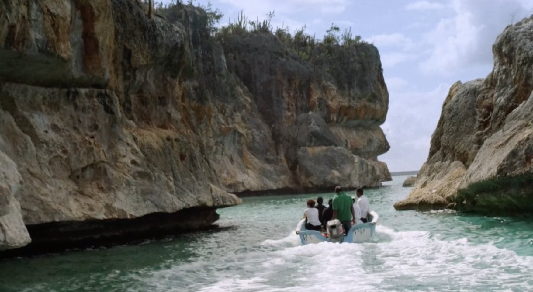 A still from a film. The image shows people in a boat sailing through some rocks.