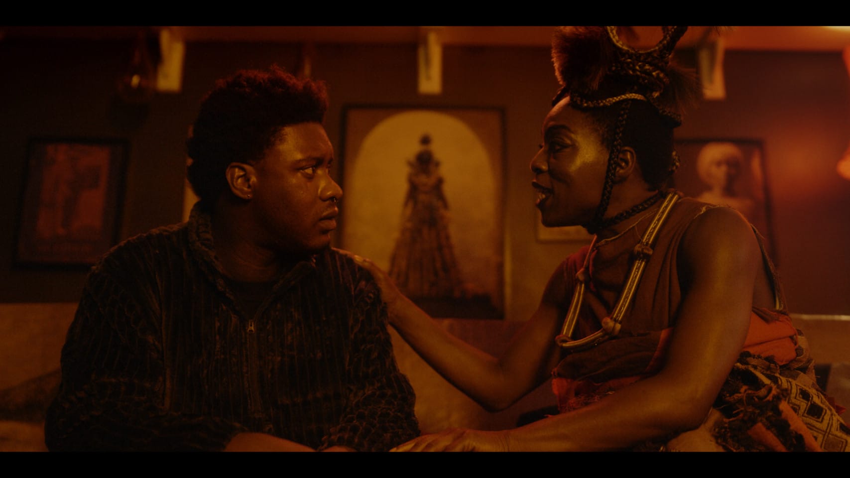 A still from a film. Two people sit together in a dark room with golden light. One person puts their hands on the other.