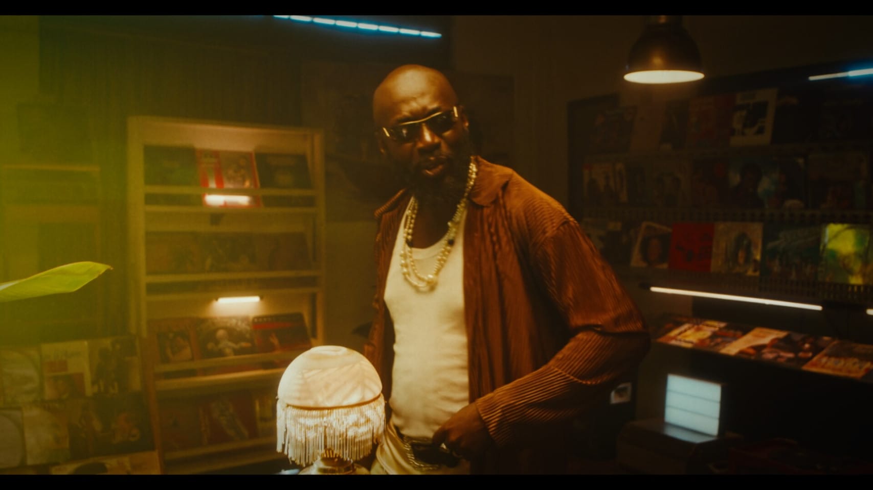 A man wearing sunglasses and necklaces stands in a dark room with his hands on his hips