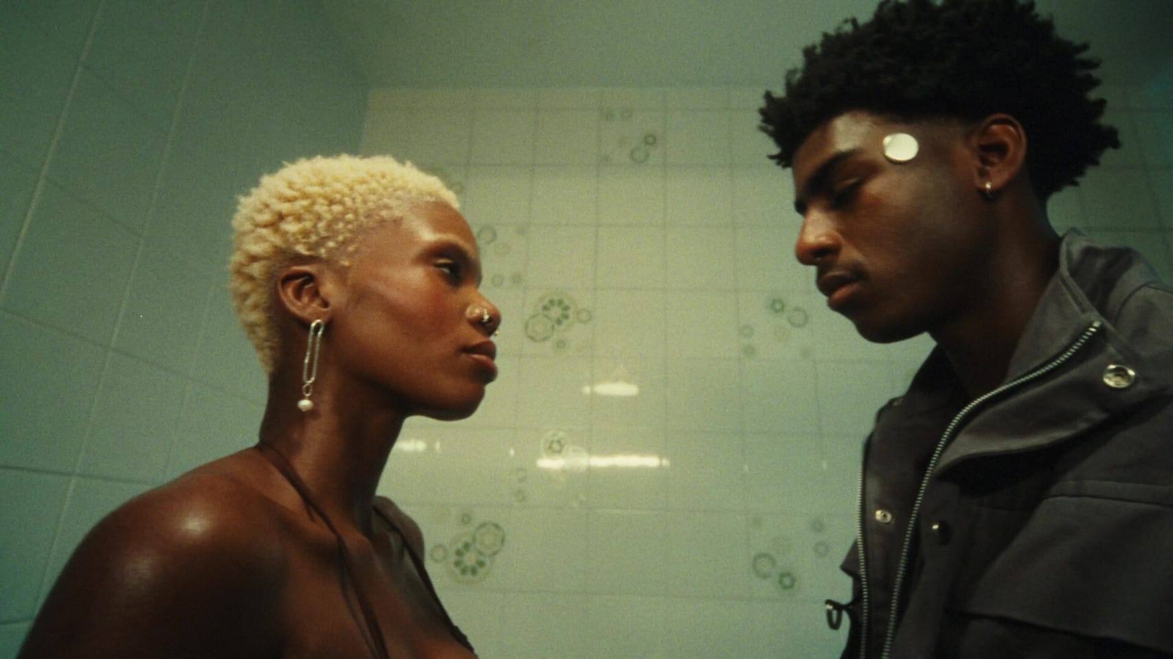 Two people stand in a tiled bathroom looking at each other with serious expressions on their faces.