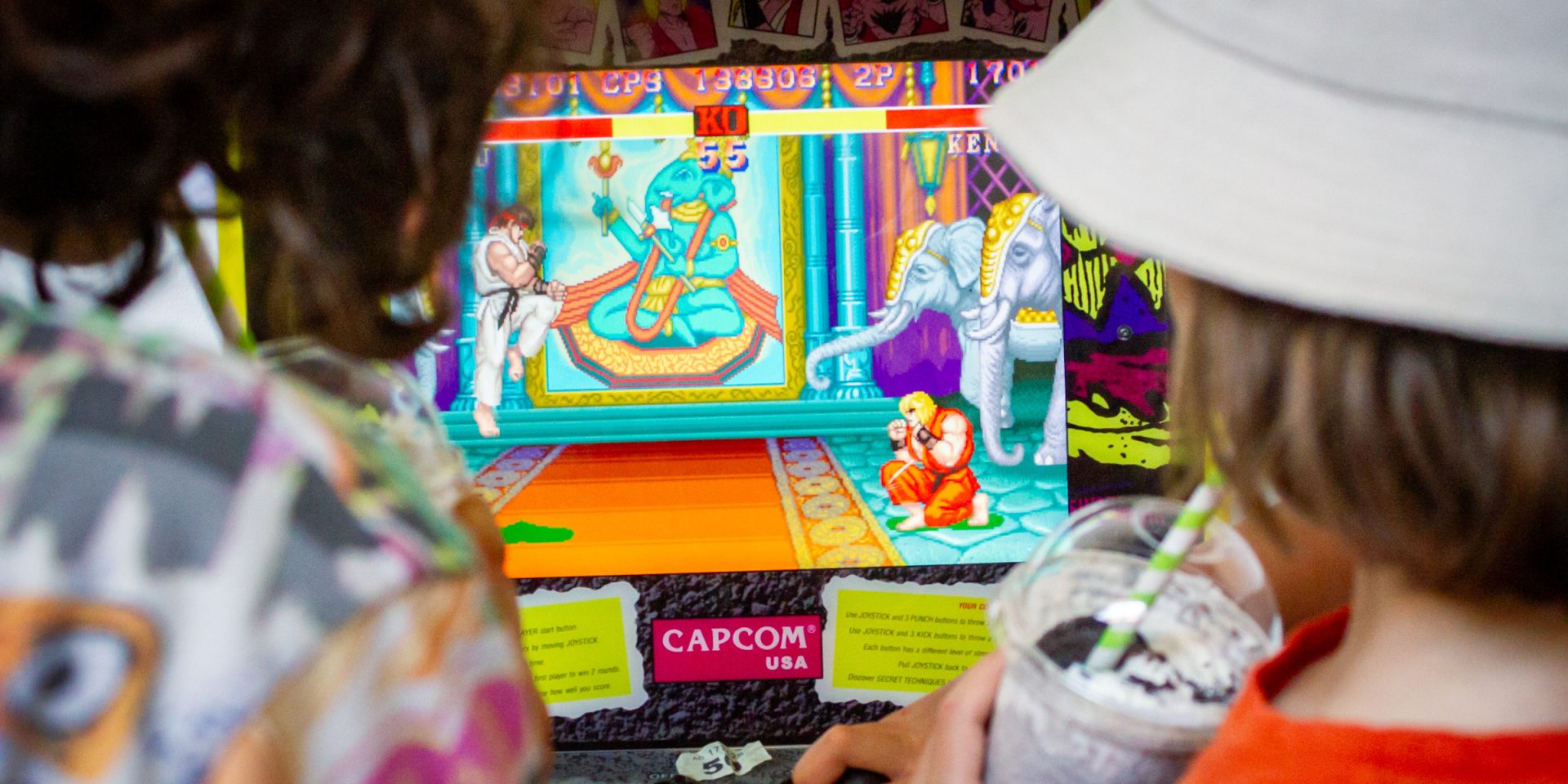 Two kids play a video game. One of the young people is wearing a white bucket hat and drinking a milkshake.
