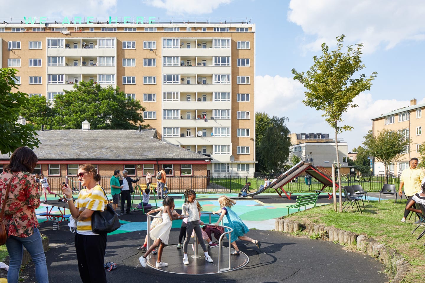 Children play on a playground in front of a large apartment building. Adults stand to the side talking.