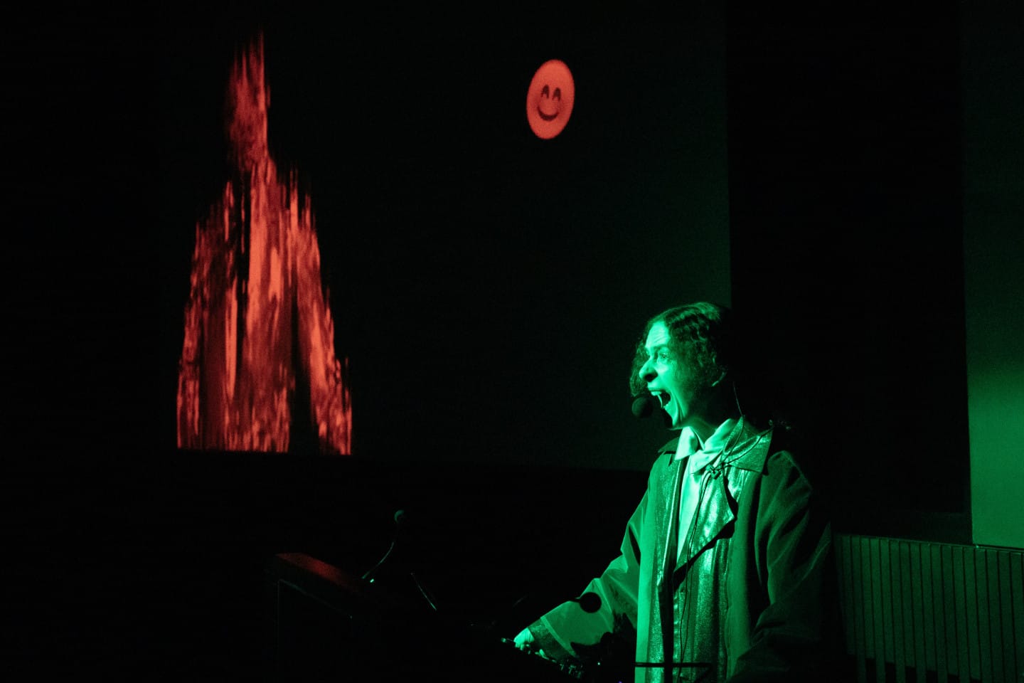 A dark room with a person screaming into a microphone as part of a performance. There is a green spotlight on the person. In the background, red projections show a smiley face and flames.