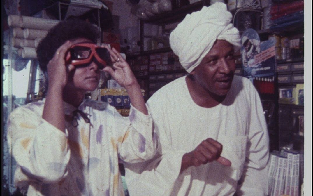 A still from a film showing two people wearing white garments in what looks like a storage room, with lots of shelves. One of them puts goggles up to their face.