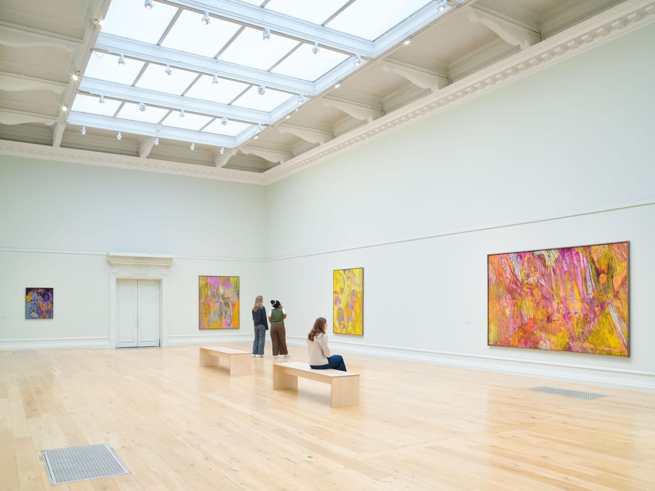 People sit and stand in a gallery and look at artworks on a wall. The paintings are by artist Christina Kimeze.