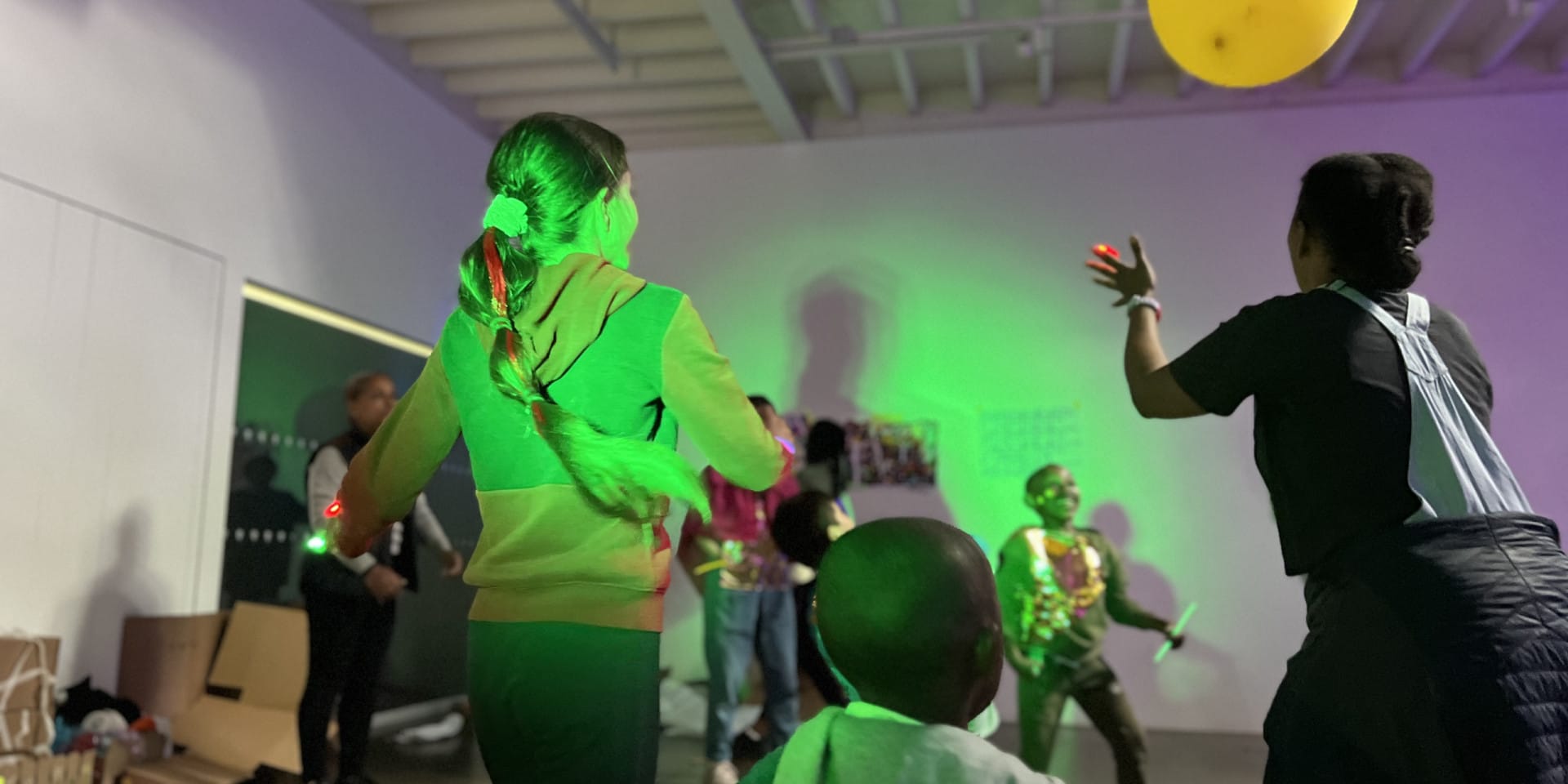 A group of children play in a large room. There are green lights shining on them and they are running around and throwing a yellow balloon.