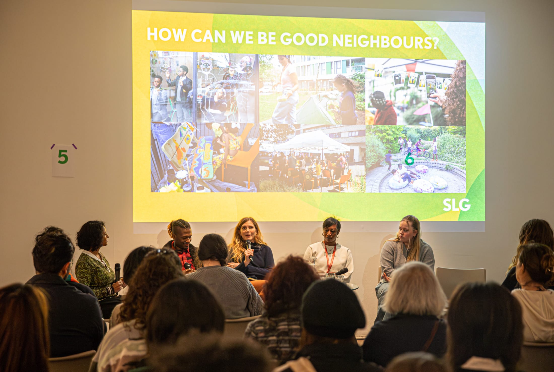 People speaking at a panel discussion. There is a projection on screen with a collage of photos. The title on the slide reads: how can we be good neighbours?