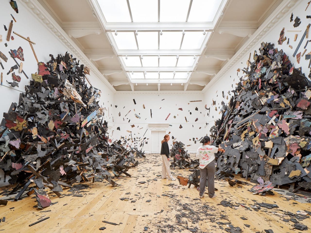 People look at a large immersive installation in the South London Gallery by artist Leonardo Drew. The work is a large explosive sculpture, with two mountainous structures crawling up two gallery walls. There is wood and debris all over the ground.