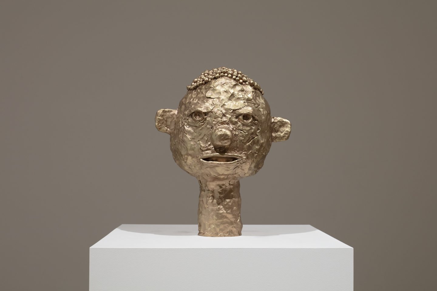 A gold sculpture of a simplified head with a gaping mouth, large nose, bullet-hole eyes, and stylized hair