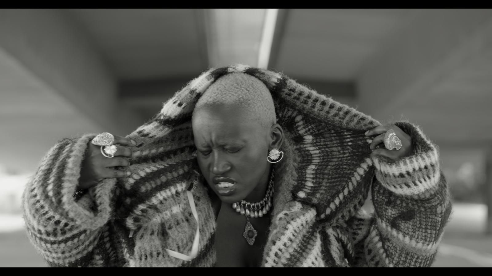 A black and white still from a music video. A woman with a shaved head wearing a knitted hoody pulls it over her head.