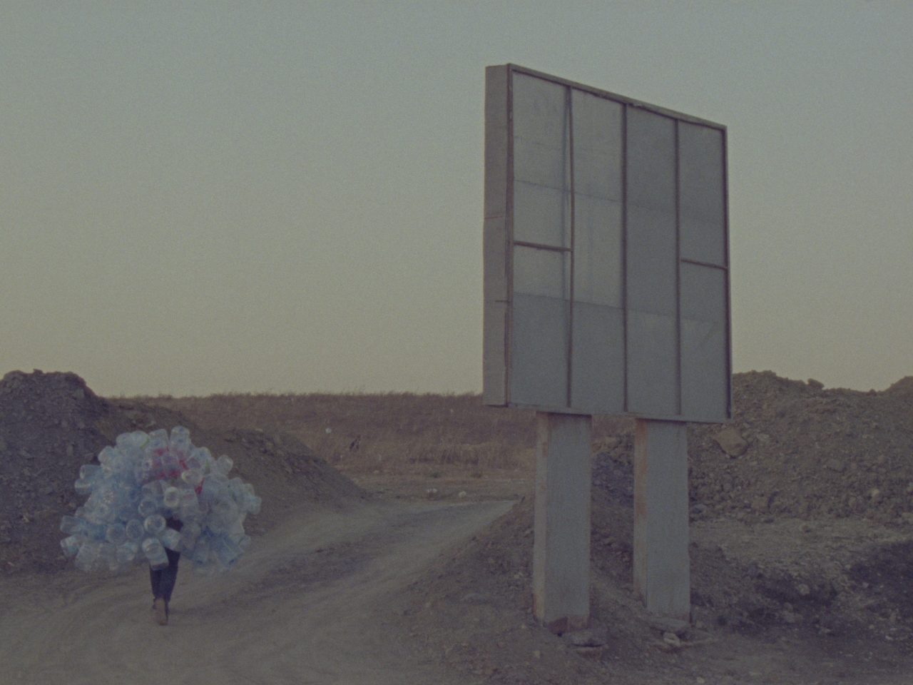A person walks through a desert-like landscape. Beside them is a billboard. They are holding lots of plastic bottles.