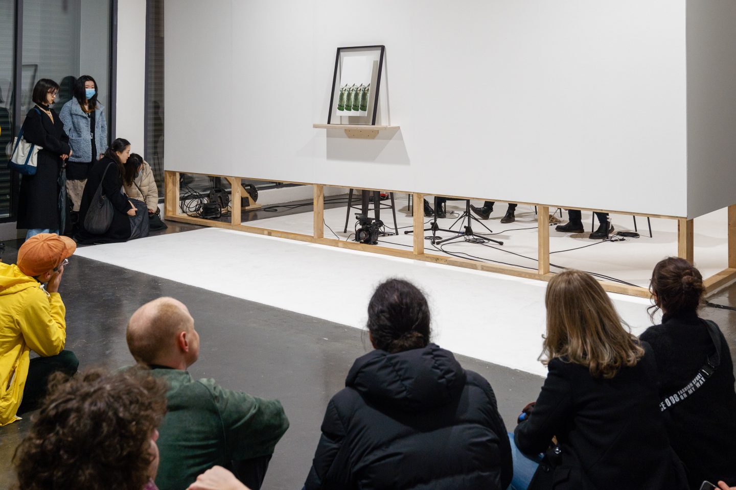 People sit around an art gallery and engage with a performance art piece. The performers are hidden behind a screen, you can only see their legs. On the screen, is an art work balancing on a shelf.